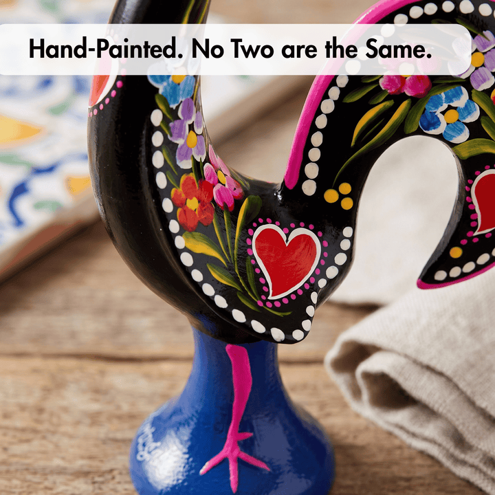 A close-up of a hand-painted portuguese rooster figurine with a glossy black body, colorful floral patterns, a red heart, and a blue base, set on a wooden surface with a text overlay reading &