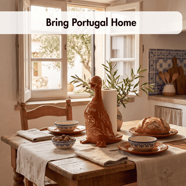 A rustic dining table set in a warm kitchen or dining room, featuring a terracotta-colored ceramic duck-shaped holder with a roll of white paper towels, blue-and-white patterned bowls, a loaf of bread, and a table runner. A white banner at the top reads &