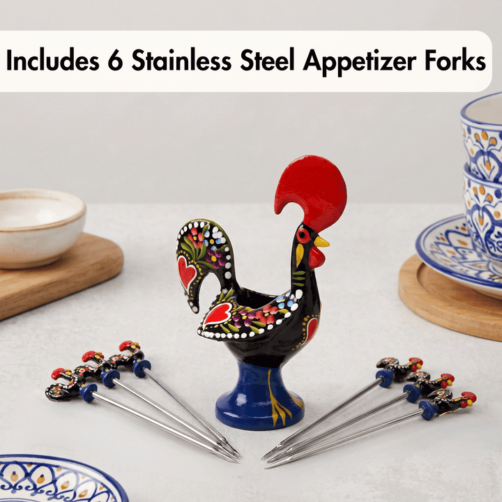 A decorative rooster-shaped holder for stainless steel appetizer forks, shown with six metal forks arranged around it on a light tabletop. The rooster has a black body with colorful hand-painted floral details, a bright red comb, and a blue base.
