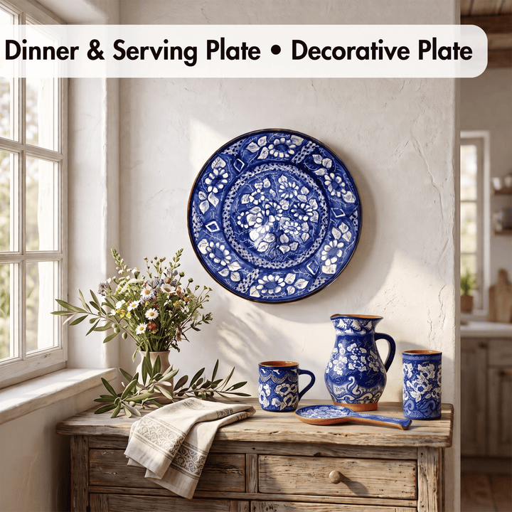 A blue and white decorative ceramic dinner plate with intricate floral patterns hangs on a textured white wall above a rustic wooden sideboard. The scene is styled with a matching blue and white pitcher, two cups, a spoon, and a vase of flowers in a bright, sunlit room.