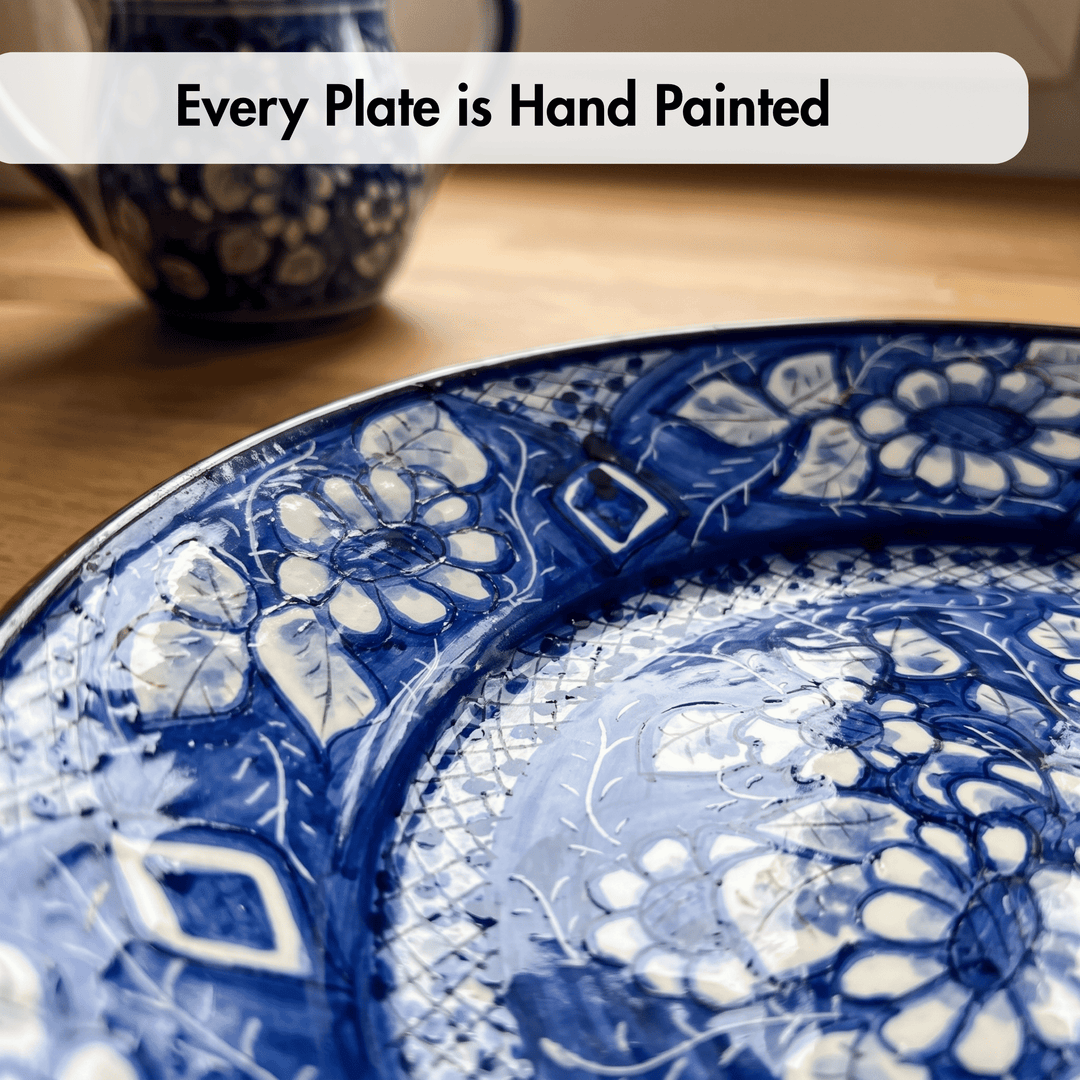 A close-up of a hand-painted ceramic plate with a glossy cobalt blue and white floral pattern, resting on a wooden surface. A blurred matching blue and white cup is visible in the background, and a text overlay at the top reads &