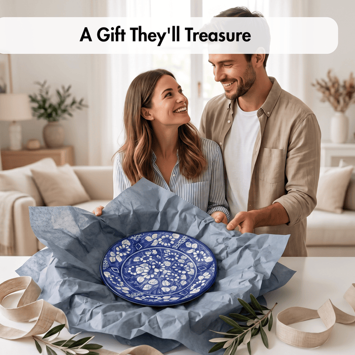 A smiling couple in a bright living room presents a blue and white floral ceramic plate wrapped in blue tissue paper. A white banner at the top reads &