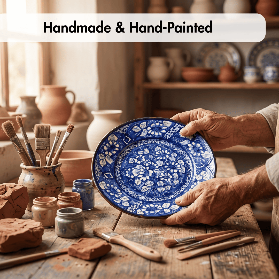 A person holds a round hand-painted ceramic plate with intricate blue floral patterns on a rustic wooden table in a pottery studio. Paintbrushes, clay, and other pottery are visible in the background, with a text banner at the top reading &