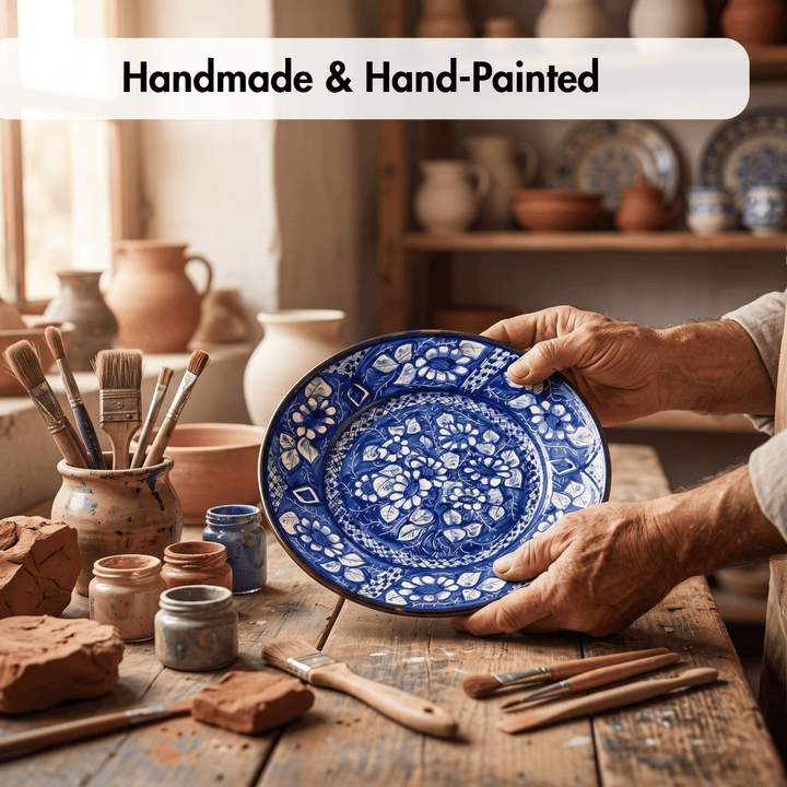 A person holds a round hand-painted ceramic plate with intricate blue floral patterns on a rustic wooden table in a pottery studio. Paintbrushes, clay, and other pottery are visible in the background, with a text banner at the top reading &