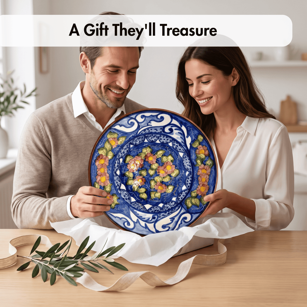 A man and a woman smile while holding a large round ceramic plate with a vibrant blue floral pattern and yellow, orange, and green accents. They are standing behind white wrapping paper on a wooden table with an olive branch in the foreground, under a text banner that reads &