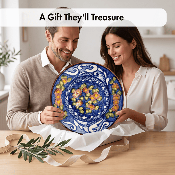A man and a woman smile while holding a large round ceramic plate with a vibrant blue floral pattern and yellow, orange, and green accents. They are standing behind white wrapping paper on a wooden table with an olive branch in the foreground, under a text banner that reads &
