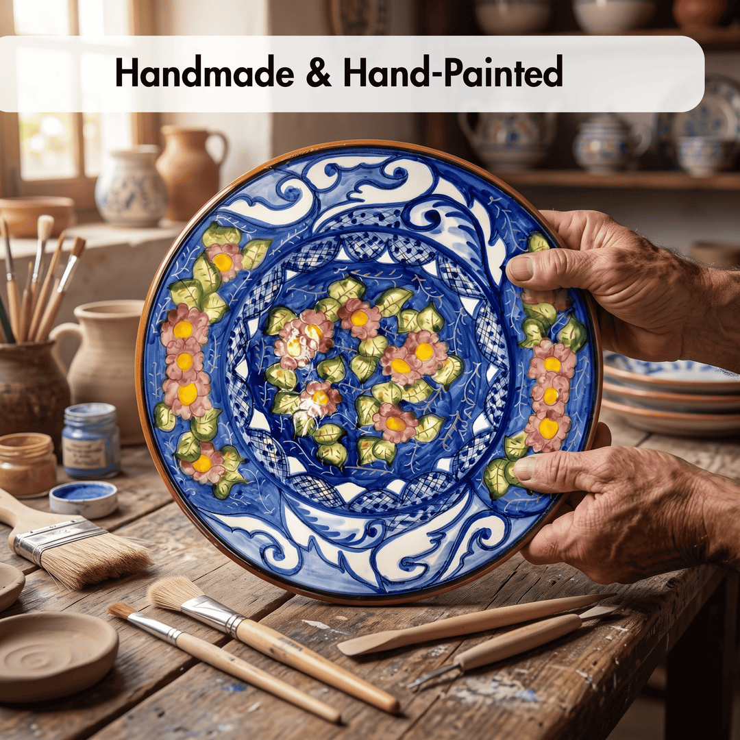 A hand-painted ceramic plate with intricate blue, white, green, and yellow floral patterns, held by hands over a rustic pottery workshop table with paintbrushes and clay jars.