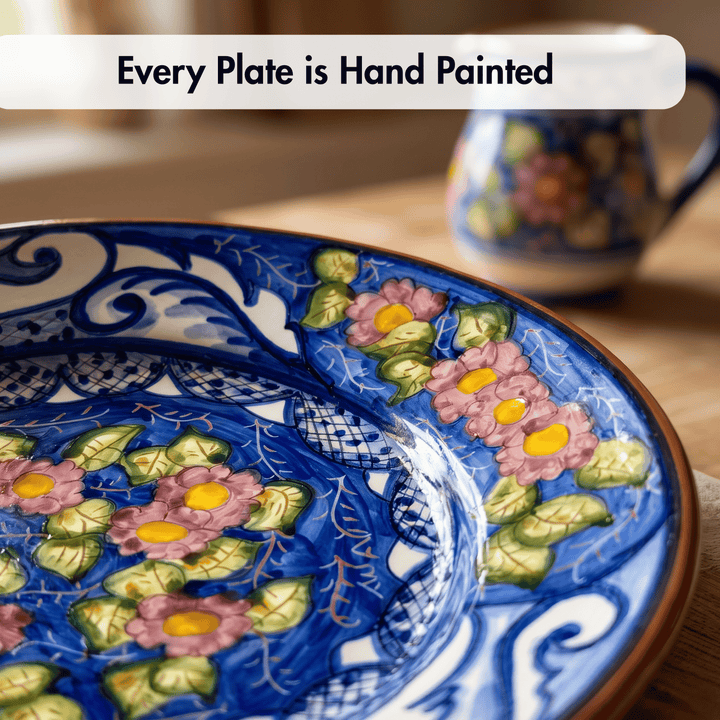 A close-up of a hand-painted ceramic plate with a blue floral pattern, pink flowers, green leaves, and yellow centers, with a blurred matching vessel in the background and the text &