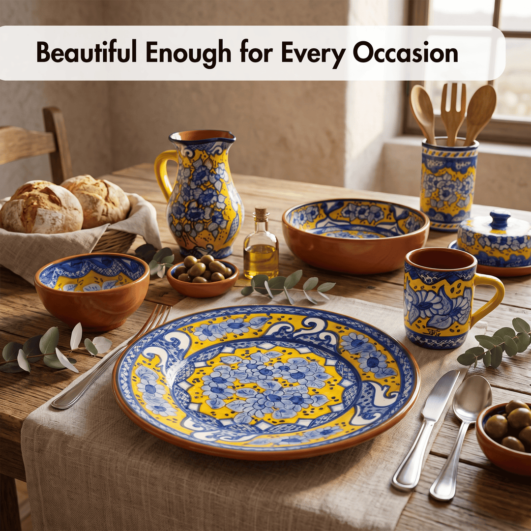 A styled table setting featuring a set of hand-painted ceramic tableware in terracotta orange with blue and yellow floral patterns. The set includes a large plate, bowls, a pitcher, a mug, utensils, and a small bottle, arranged on a linen table runner with bread, olives, and eucalyptus leaves.