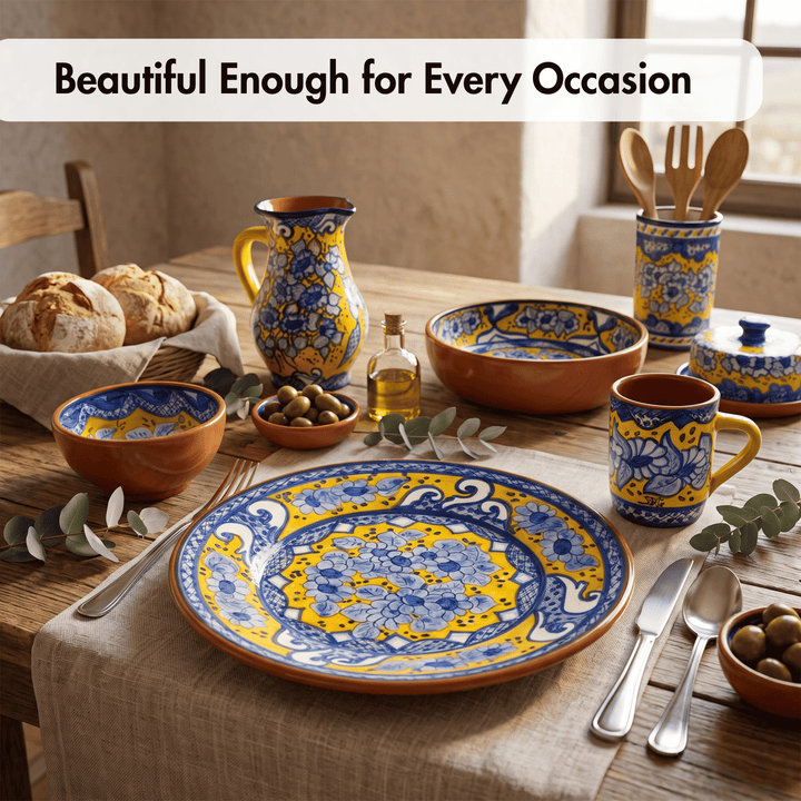 A styled table setting featuring a set of hand-painted ceramic tableware in terracotta orange with blue and yellow floral patterns. The set includes a large plate, bowls, a pitcher, a mug, utensils, and a small bottle, arranged on a linen table runner with bread, olives, and eucalyptus leaves.