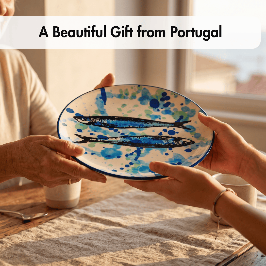 A ceramic oval plate with blue painted fish and watercolor-style blue splatters on a white base, held by two hands over a table in a warm indoor setting. Text at the top reads &