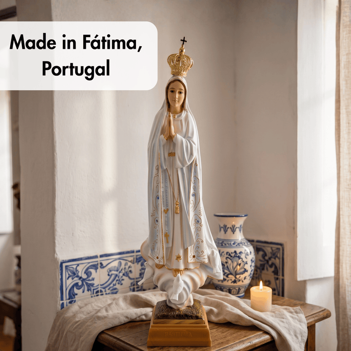 A religious statue of the Virgin Mary wearing a white robe with gold detailing and a gold crown, displayed on a wooden table in a softly lit room. A blue-and-white ceramic vase, a lit candle, and a white text box reading &