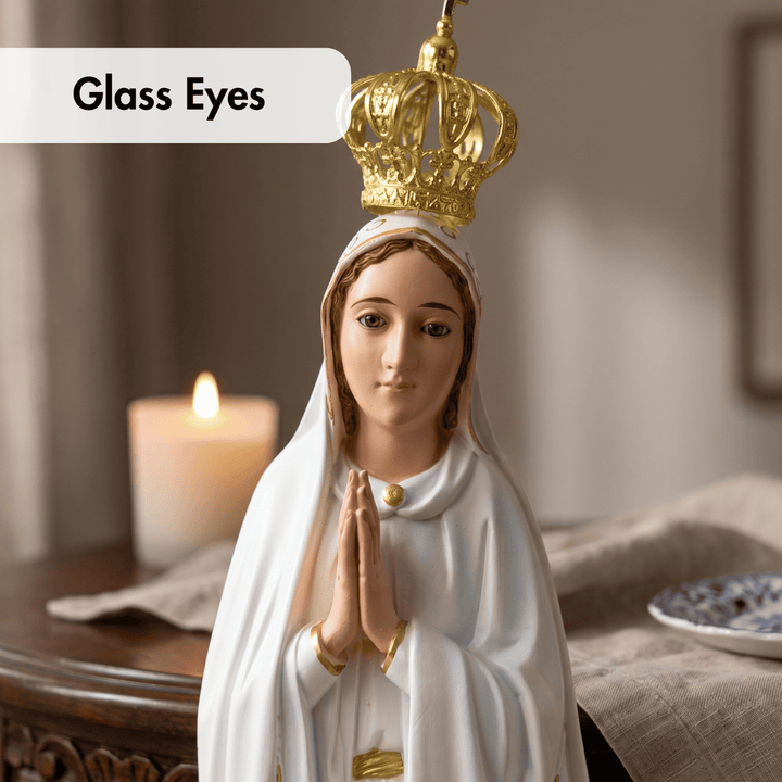 A white and gold religious figurine of the Virgin Mary with clasped hands and a crown, shown in a softly lit indoor setting with a candle in the background. A label in the top left reads &