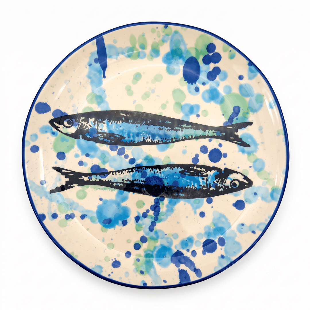 A round ceramic plate with a hand-painted design of two blue and black fish on a cream background with blue and green watercolor splatters, finished with a dark blue rim.