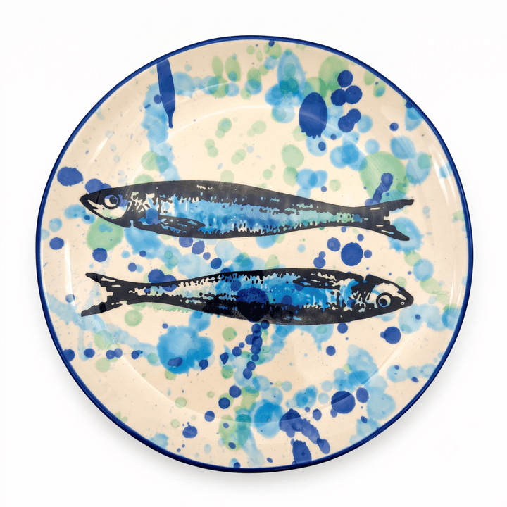 A round ceramic plate with a hand-painted design of two blue and black fish on a cream background with blue and green watercolor splatters, finished with a dark blue rim.
