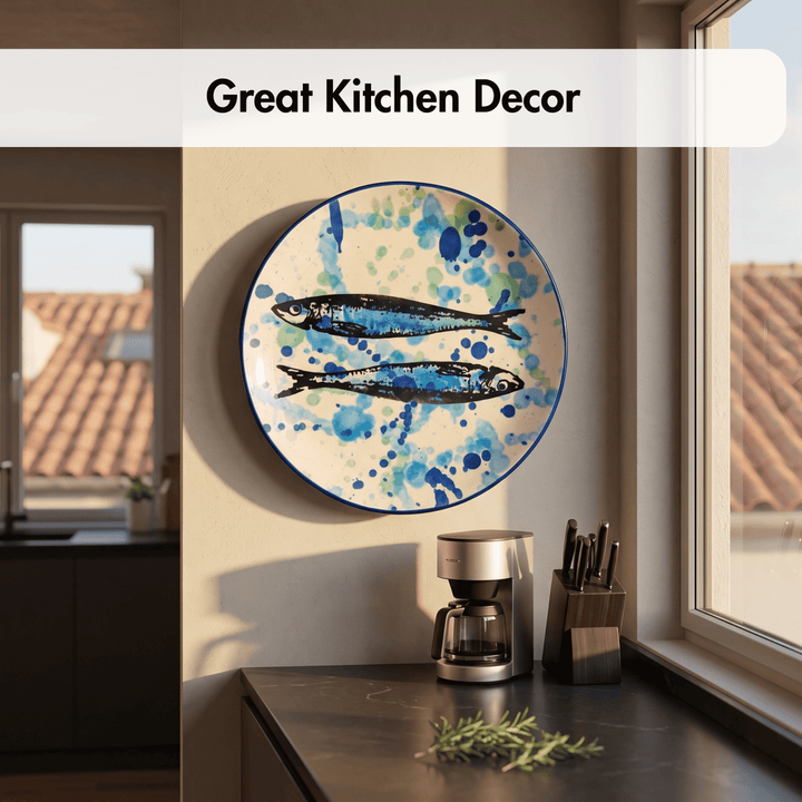 A round decorative wall plate with two blue fish illustrations and blue watercolor splatters on a cream background, displayed in a kitchen setting above a countertop with a coffee maker.