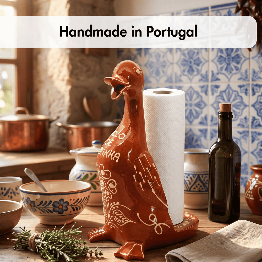 A brown ceramic duck-shaped paper towel holder with white painted floral details, holding a roll of white paper towels. The scene is set on a rustic kitchen counter with blue and white tiles, copper pots, a dark glass bottle, and a sprig of rosemary in the foreground.