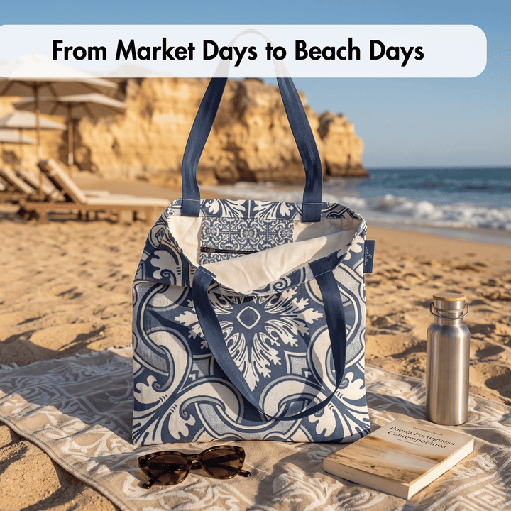 A blue and white patterned tote bag sits on a beach towel on the sand, with sunglasses, a book, and a metal water bottle beside it. The background shows a beach scene with ocean waves and rock formations.