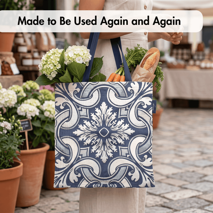 A person at an outdoor market holds a reusable tote bag with a navy and white ornate tile pattern. The bag is filled with flowers, carrots, and a loaf of bread.