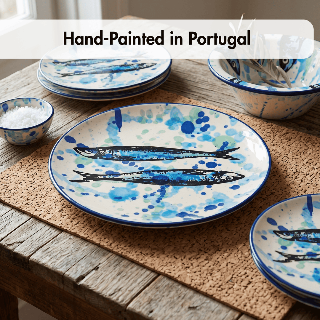 A set of hand-painted ceramic tableware featuring blue and black fish designs on a white background, displayed on a rustic wooden table with a woven placemat. A text overlay at the top reads &