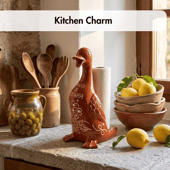 A terracotta-colored ceramic duck-shaped paper towel holder with white painted details sits on a stone kitchen counter next to lemons, bowls, wooden utensils, and a jar of olives. A white banner at the top reads &
