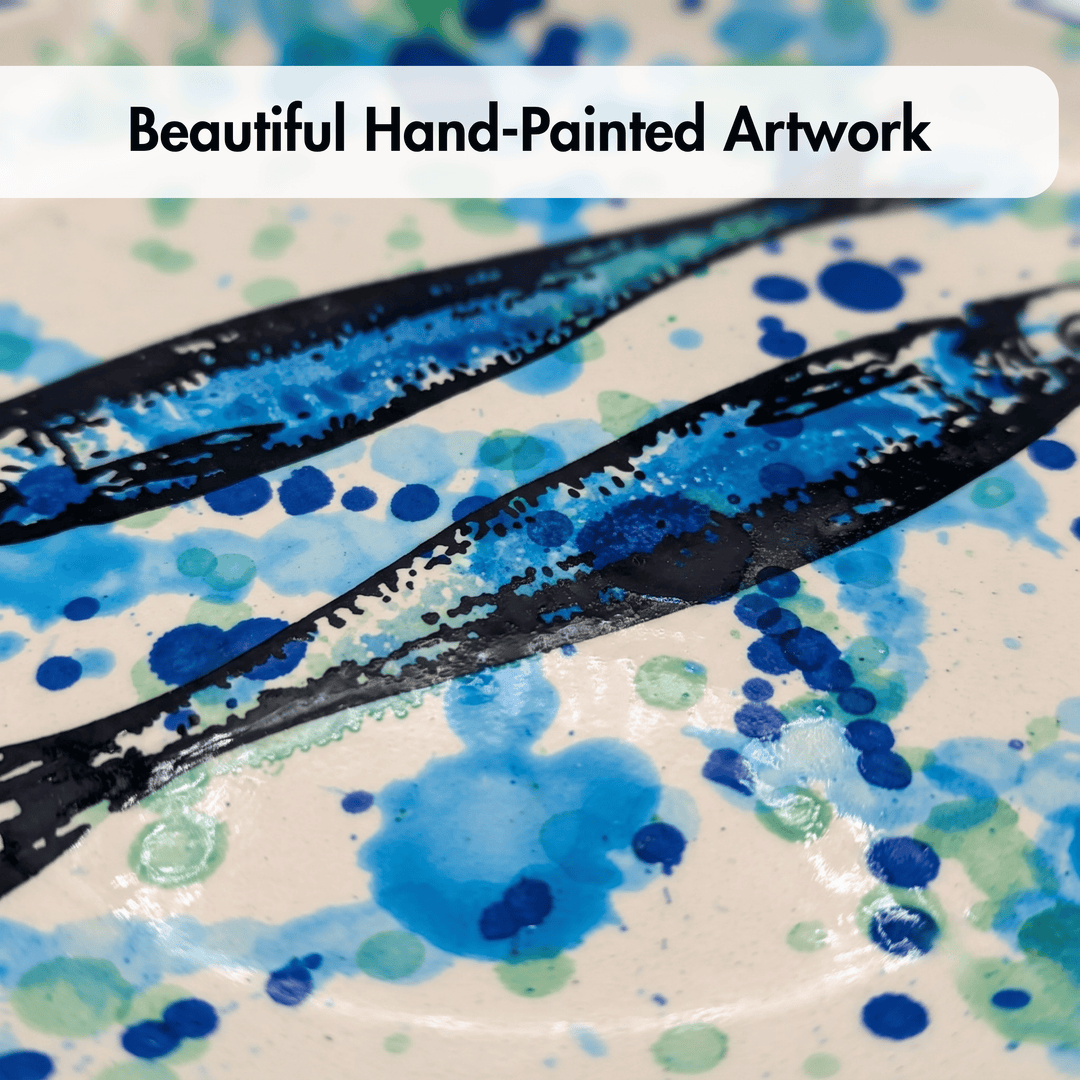 Close-up of a hand-painted ceramic plate featuring two black fish silhouettes filled with blue and green watercolor splatters on a cream background, with a white banner at the top reading &