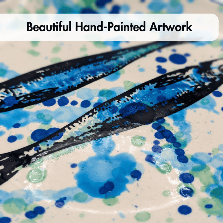 Close-up of a hand-painted ceramic plate featuring two black fish silhouettes filled with blue and green watercolor splatters on a cream background, with a white banner at the top reading &