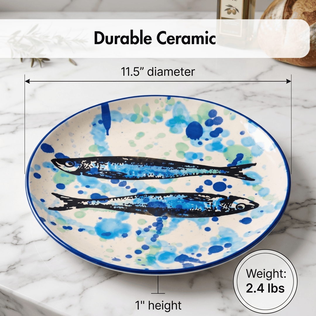 A ceramic serving platter with two painted blue fish on a white background with blue and green watercolor splatters, shown on a marble surface with dimension lines and text reading &
