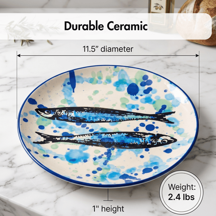 A ceramic serving platter with two painted blue fish on a white background with blue and green watercolor splatters, shown on a marble surface with dimension lines and text reading &