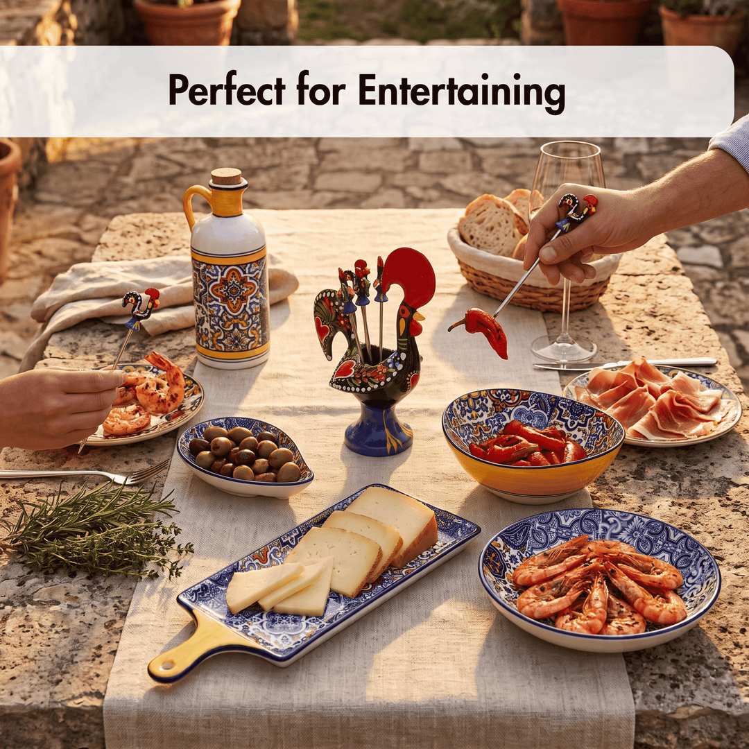 A styled outdoor dining table set with colorful ceramic serving dishes, bread, shrimp, olives, sliced cheese, cured meat, and a decorative rooster-shaped charcuterie stick holder. A hand is holding a red pepper on a metal spear, and another hand is reaching for food.