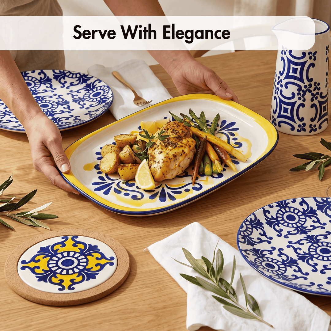 A rectangular serving platter with a yellow rim and blue floral pattern, holding cooked chicken, potatoes, lemon slices, and carrots on a wooden table. Hands are visible holding the platter, with matching blue-and-white plates, a mug, coasters, napkins, and olive branches around it.