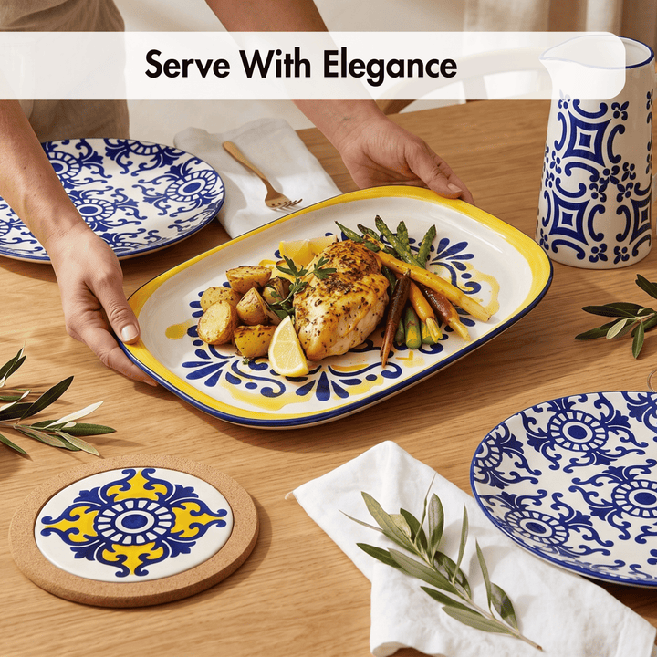A rectangular serving platter with a yellow rim and blue floral pattern, holding cooked chicken, potatoes, lemon slices, and carrots on a wooden table. Hands are visible holding the platter, with matching blue-and-white plates, a mug, coasters, napkins, and olive branches around it.