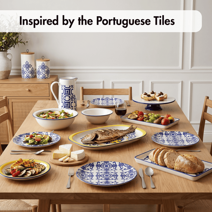 A dining table set with multiple ceramic plates, bowls, and a pitcher in a bright dining room. The tableware features a classic blue and white Portuguese tile pattern, styled with food including fish, bread, salad, and fruit.
