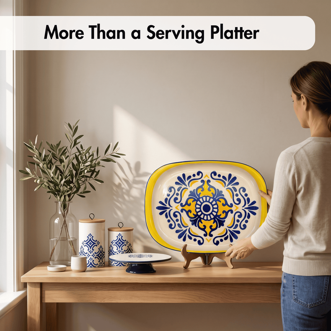 A woman in a beige sweater and blue jeans stands beside a wooden table, holding a large oval serving platter with a yellow border and blue floral tile-style pattern. The scene includes smaller matching ceramic canisters, a glass vase with greenery, and a white text banner at the top reading &