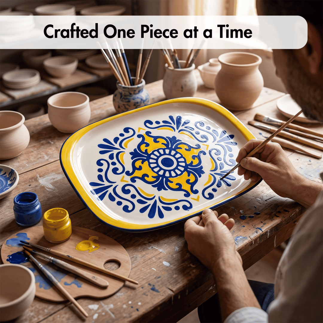 A person painting a decorative ceramic serving platter with a blue and yellow floral pattern on a wooden table surrounded by pottery tools and clay pots.