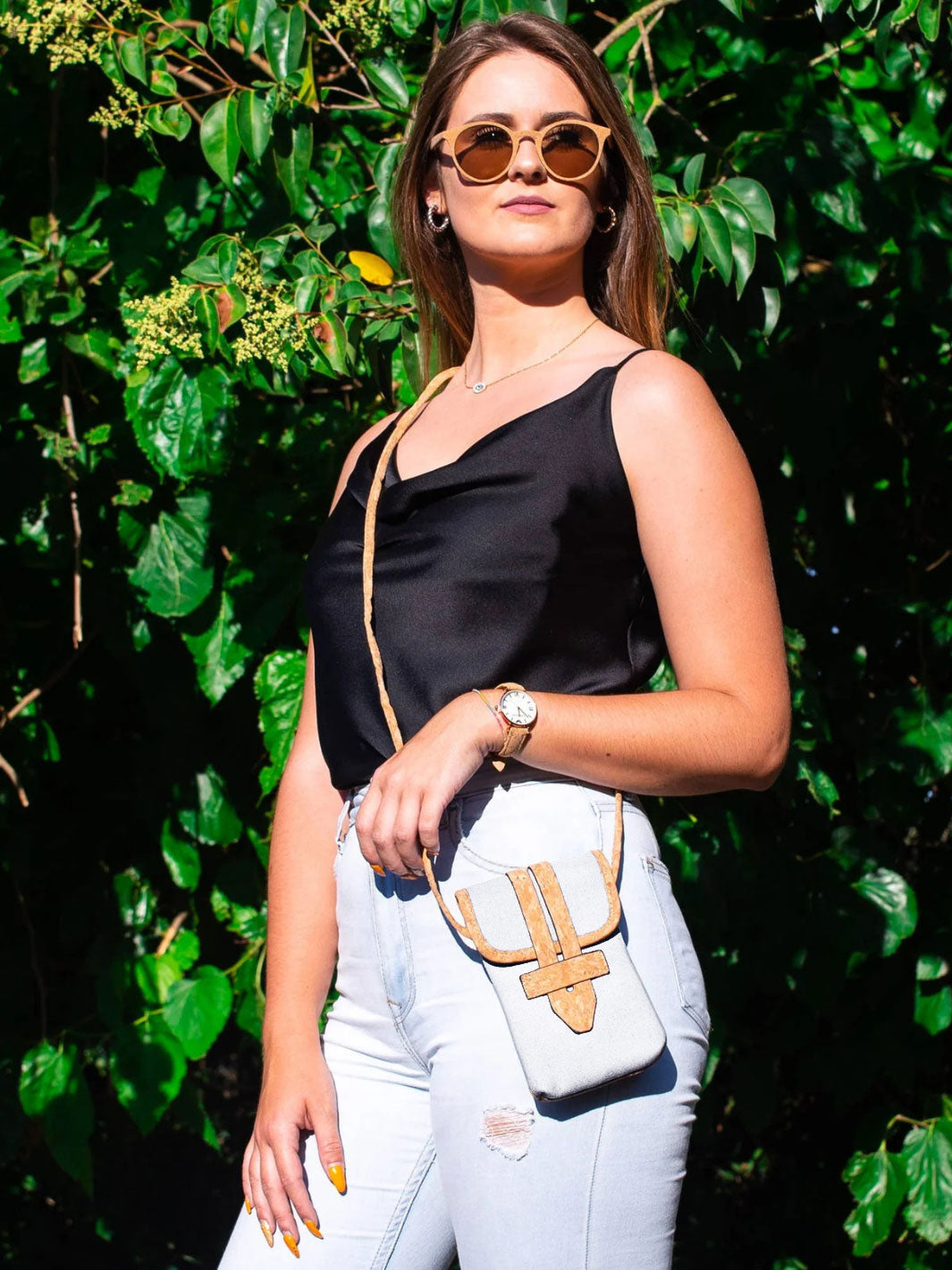 Woman wearing a black top and white pants with a light-colored bag against a green leafy background