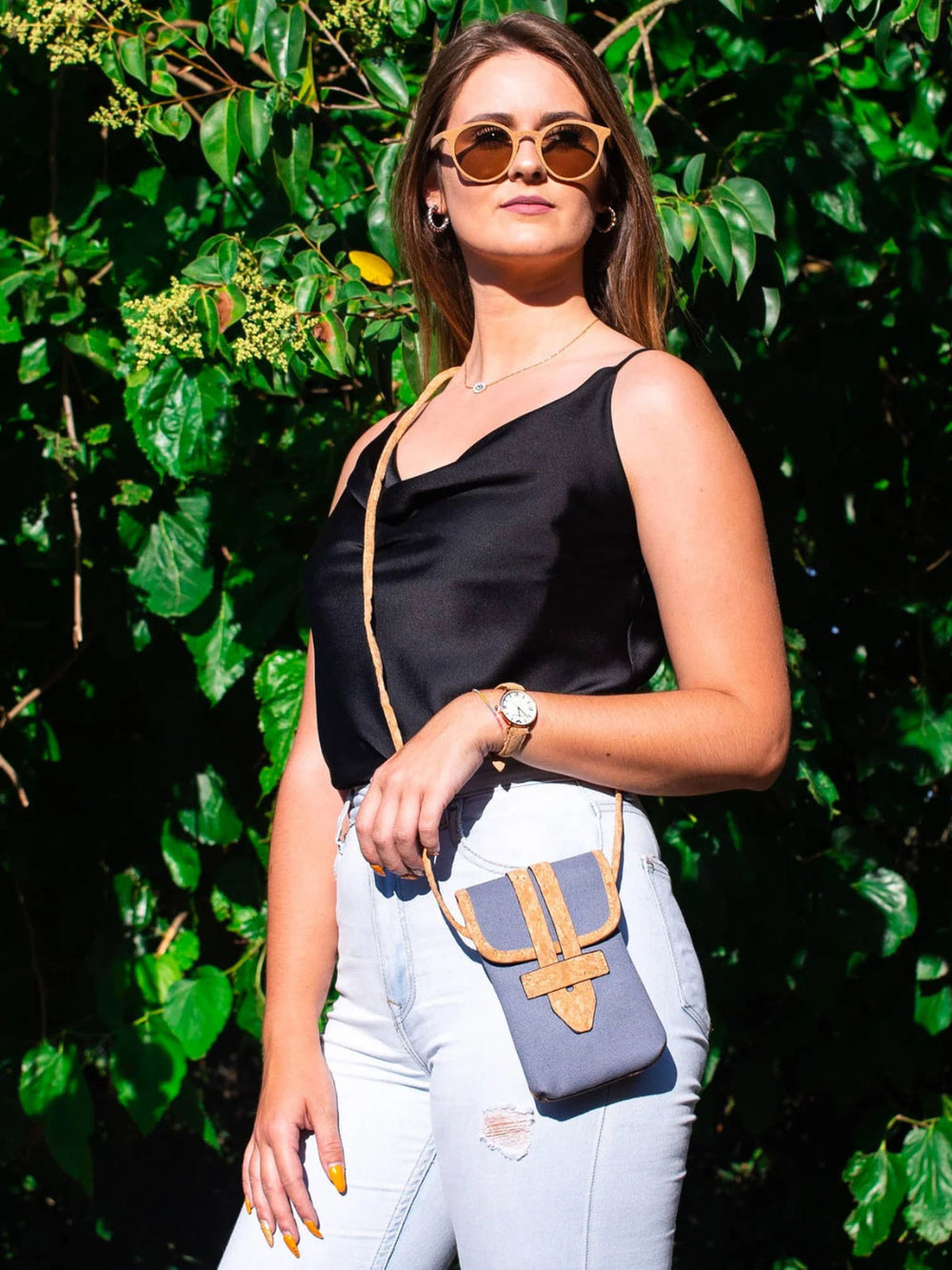 Woman wearing a black top and white pants with a blue handbag against a green leafy background