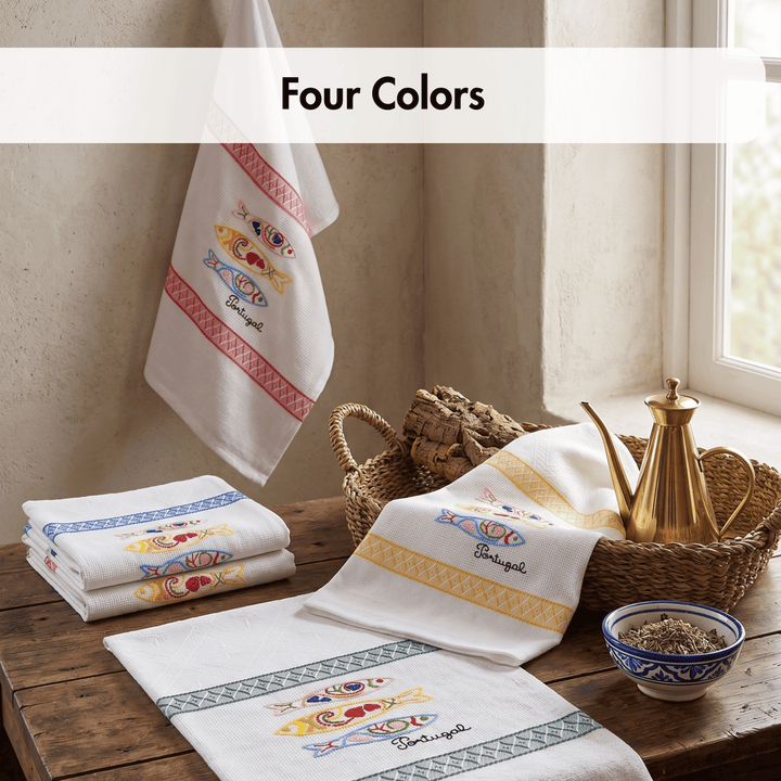Four folded and hanging white towels with colorful embroidered fish motifs and decorative borders, arranged on a rustic wooden table beside a brass teapot, wicker basket, and bowl. A banner at the top reads &