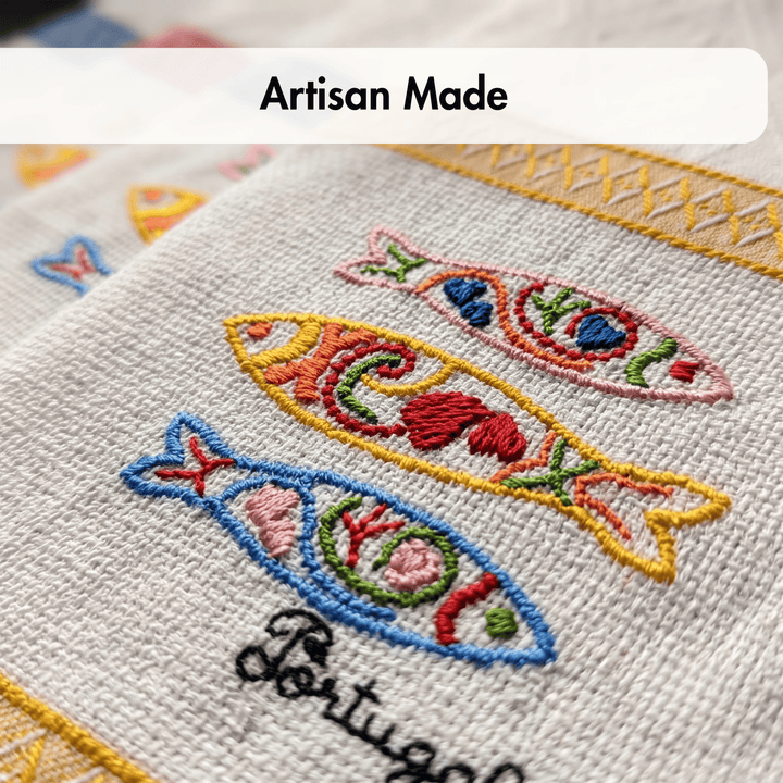 Close-up of a woven textile coaster or mat with embroidered fish motifs in pink, yellow, blue, red, and green, plus a yellow border and the word &