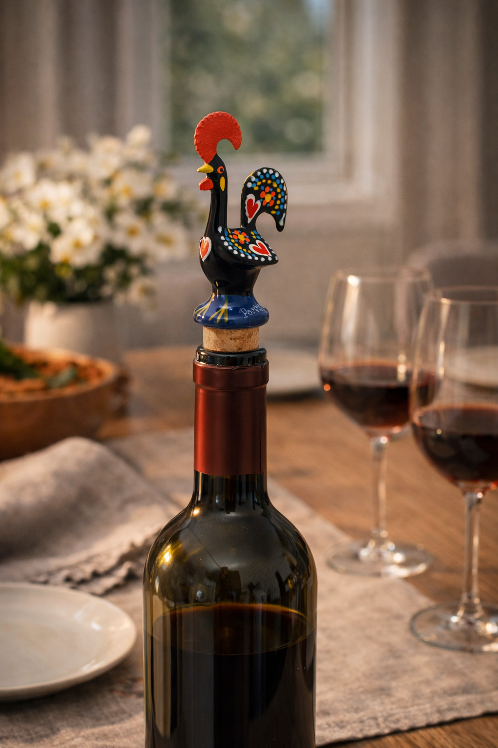 Wine bottle with decorative rooster stopper on a table with glasses of red wine and flowers.