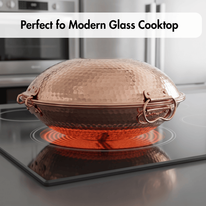 A hammered copper dome-shaped pot lid with copper handles sits on a black glass cooktop with the burner turned on, emitting a red glow. The background shows a blurred modern kitchen setting with stainless steel appliances.