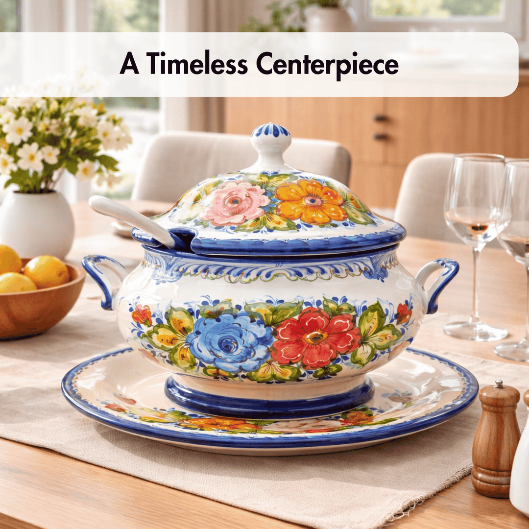 A hand-painted ceramic tureen with a matching lid and saucer, decorated with vibrant floral motifs in blue, red, pink, and yellow. It is styled on a wooden dining table with a placemat, wine glasses, and a bowl of lemons in the background, under the text overlay &
