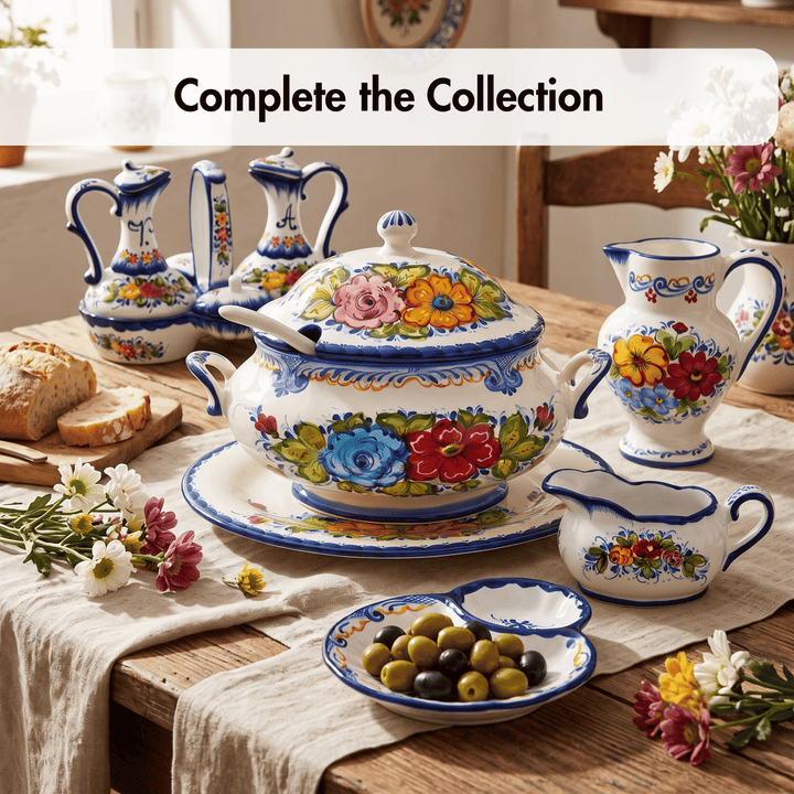 A coordinated set of hand-painted ceramic tableware with a white base and colorful floral motifs, arranged on a wooden table with bread, olives, flowers, and a cream-colored runner. Text at the top reads &