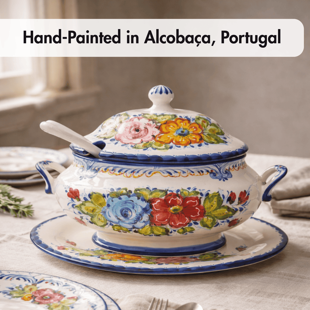 A hand-painted ceramic tureen with lid and matching saucer, decorated with colorful floral motifs and blue trim, set on a table. Text at the top reads 'Hand-Painted in Alcobaça, Portugal'.