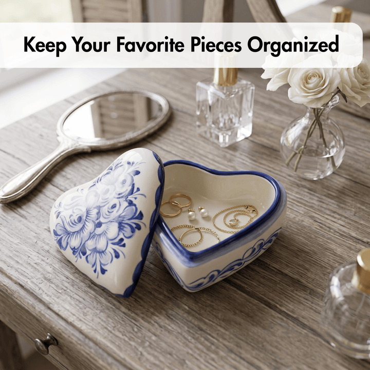 A heart-shaped ceramic jewelry box with a blue floral pattern on the lid, shown open on a wooden table with rings inside. A hand mirror, perfume bottles, and white flowers are arranged around it, with the text &