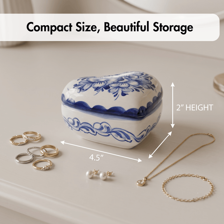 A heart-shaped white ceramic jewelry box with blue floral patterns, shown on a light beige surface with surrounding jewelry and measurement arrows labeled 4.5 inches wide and 2 inches high.