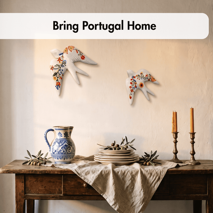 Two white ceramic birds with colorful floral patterns are mounted on a beige wall above a rustic wooden table styled with a blue-and-white ceramic pitcher, plates, candles, and olive branches. A white banner at the top reads &