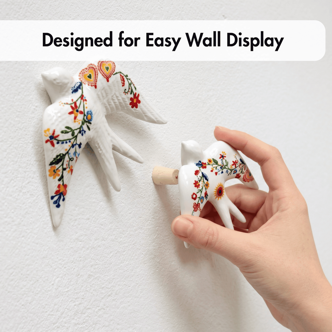 A hand is holding a small white ceramic bird wall hook with colorful floral patterns, while another matching bird hook is mounted on a white textured wall. Text at the top reads &