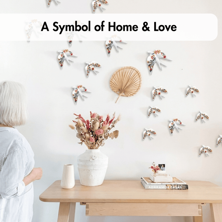 A woman with short white hair stands beside a light wood table in a bright room, looking at a wall decorated with many small white and orange floral bird cutouts and a woven round wall hanging. A white banner at the top reads, &