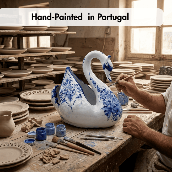 A hand-painted ceramic swan pitcher with blue floral patterns on a white glaze, being painted by a craftsman in a pottery studio. The text &